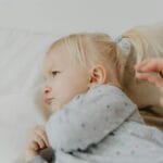 Niezadowolona dwuletnia dziewczynka z kitką siedzi naburmuszona na łóżku