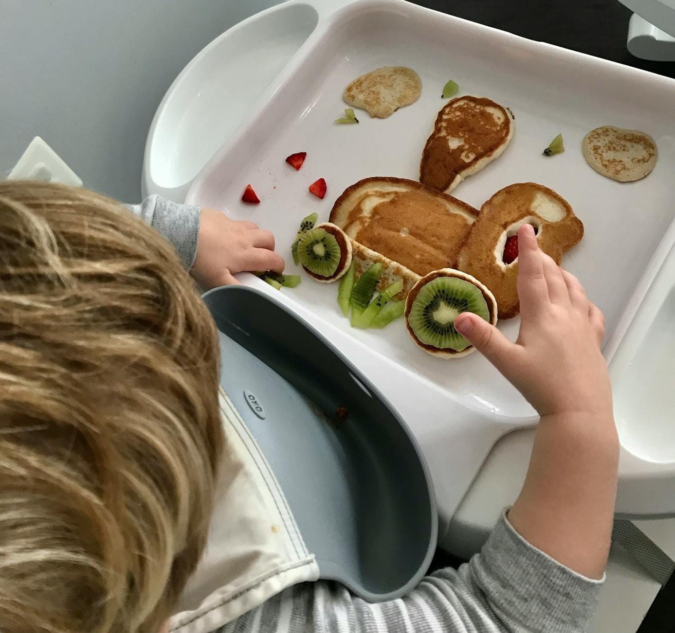 Dziecko na krzesełku tworzące śniadanie z naleśników i owoców