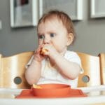 Pierwsze dni rozszerzania diety: co podać, by uniknąć problemów z brzuszkiem? Niemowlę w krzesełku do karmienia je pierwszy kawałek owocu